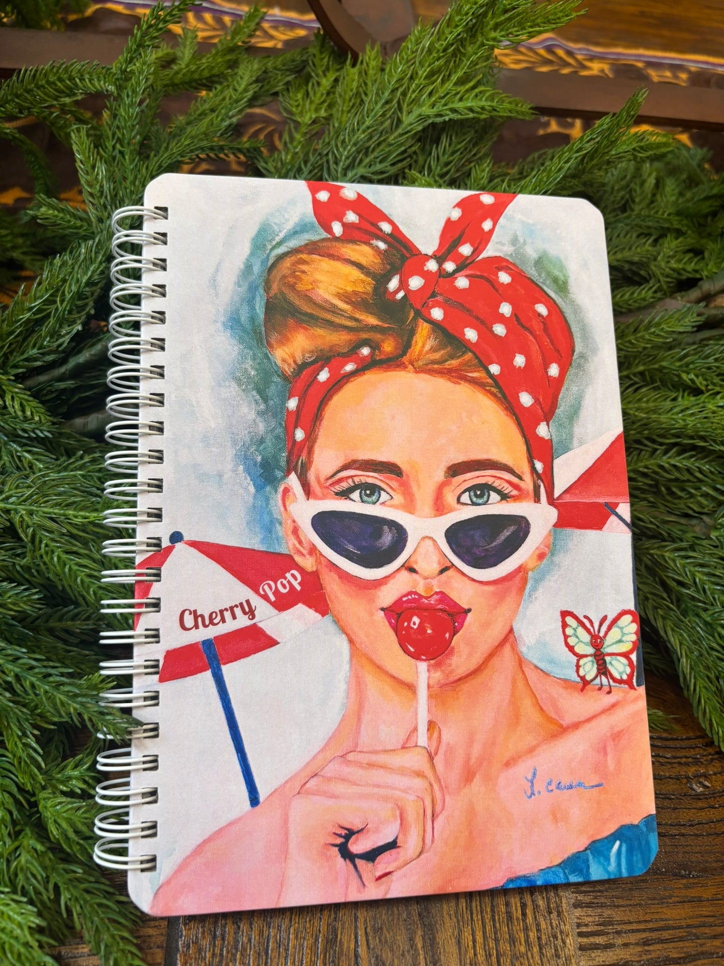 Cherry Pop art notebook featuring a retro red-haired girl with a lollipop, styled on a bed of Christmas greenery; festive colorful 60s Charmers aesthetic journal photo.