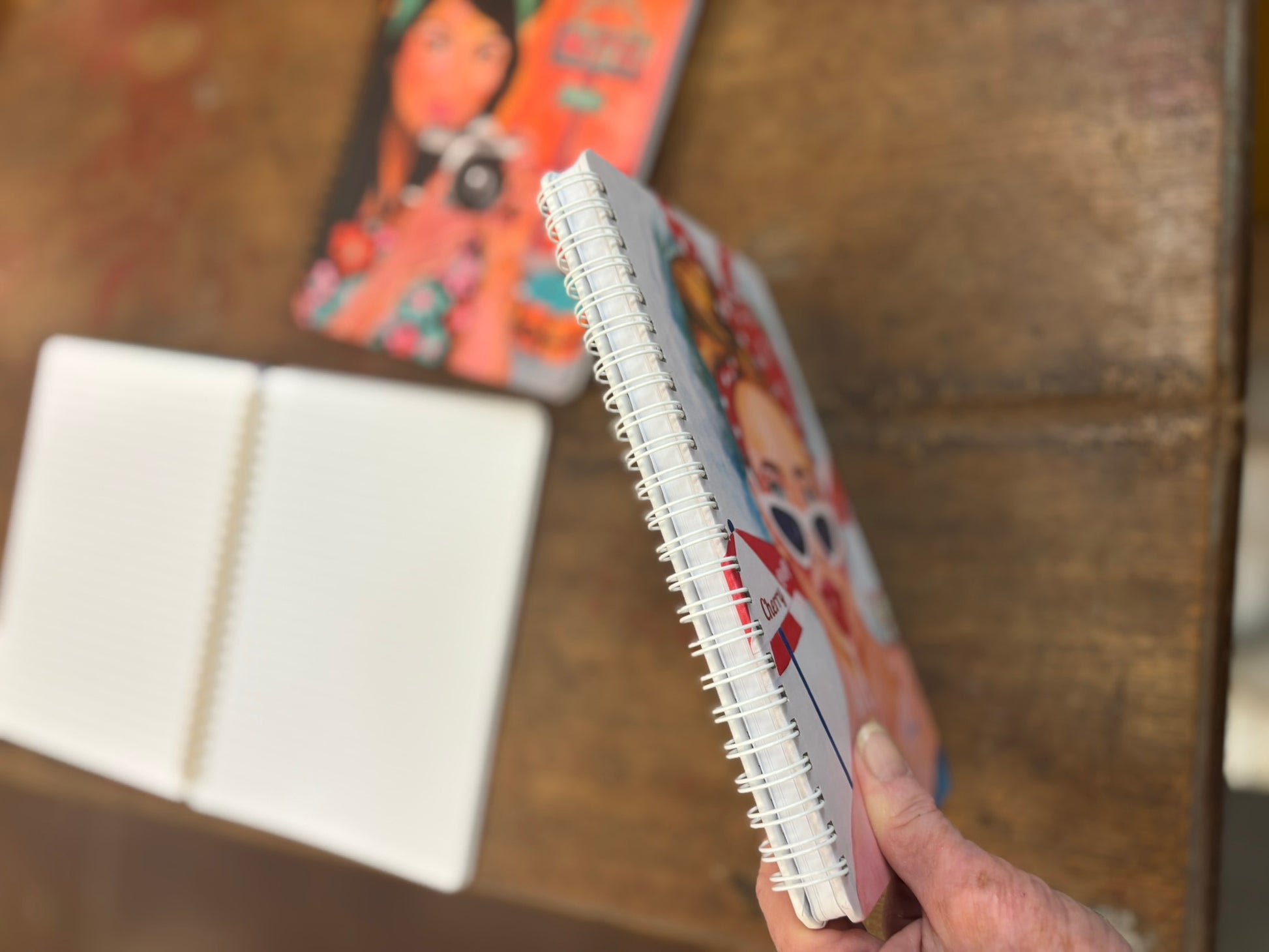 Close-up of notebook spine with an open lined journal in the background and the Shutter Bell cover blurred on the table; detail photo showing notebook quality and interior pages.