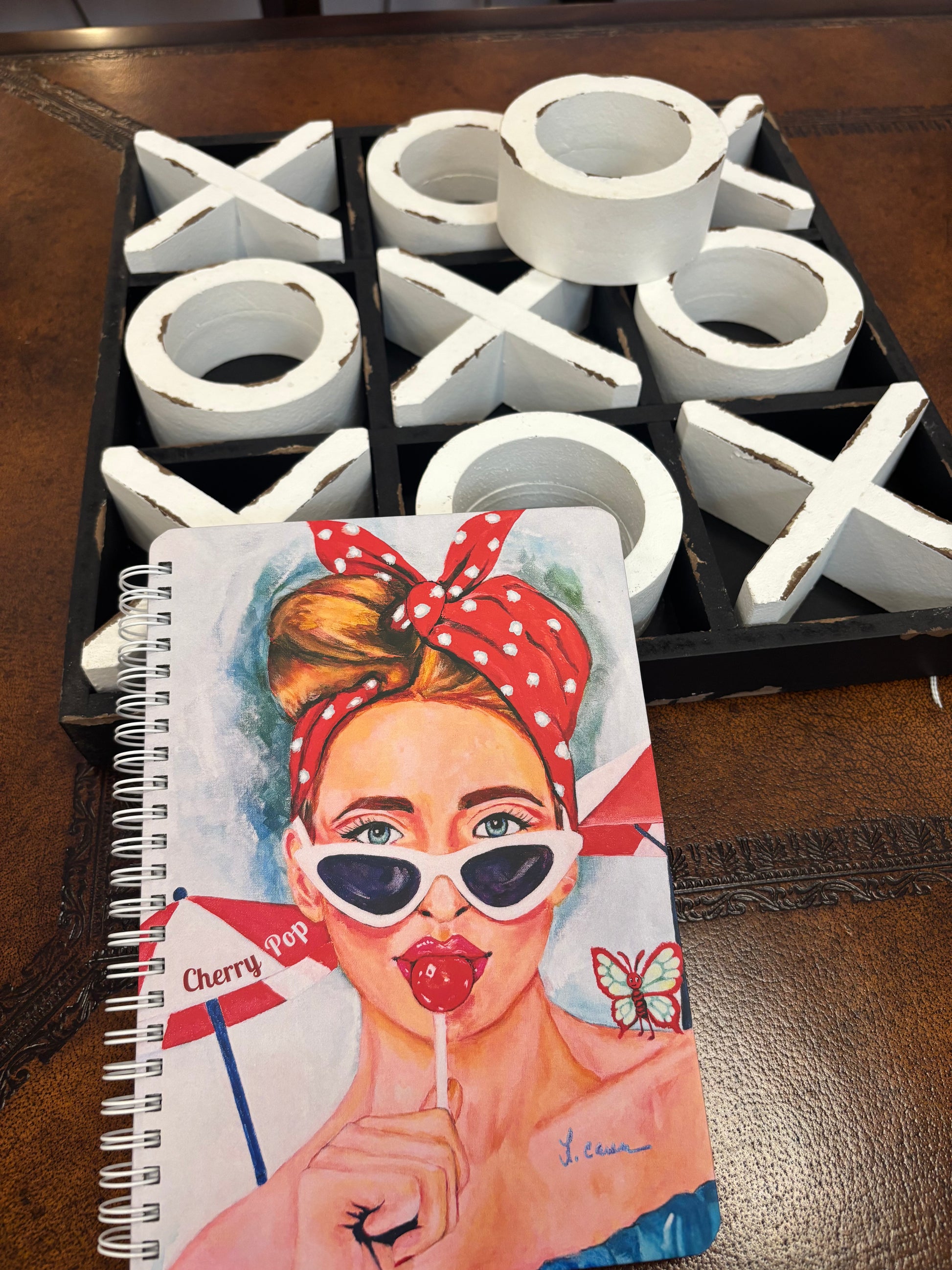 Notebook with a colorful cover featuring a woman eating a lollipop, next to a tic-tac-toe game set.