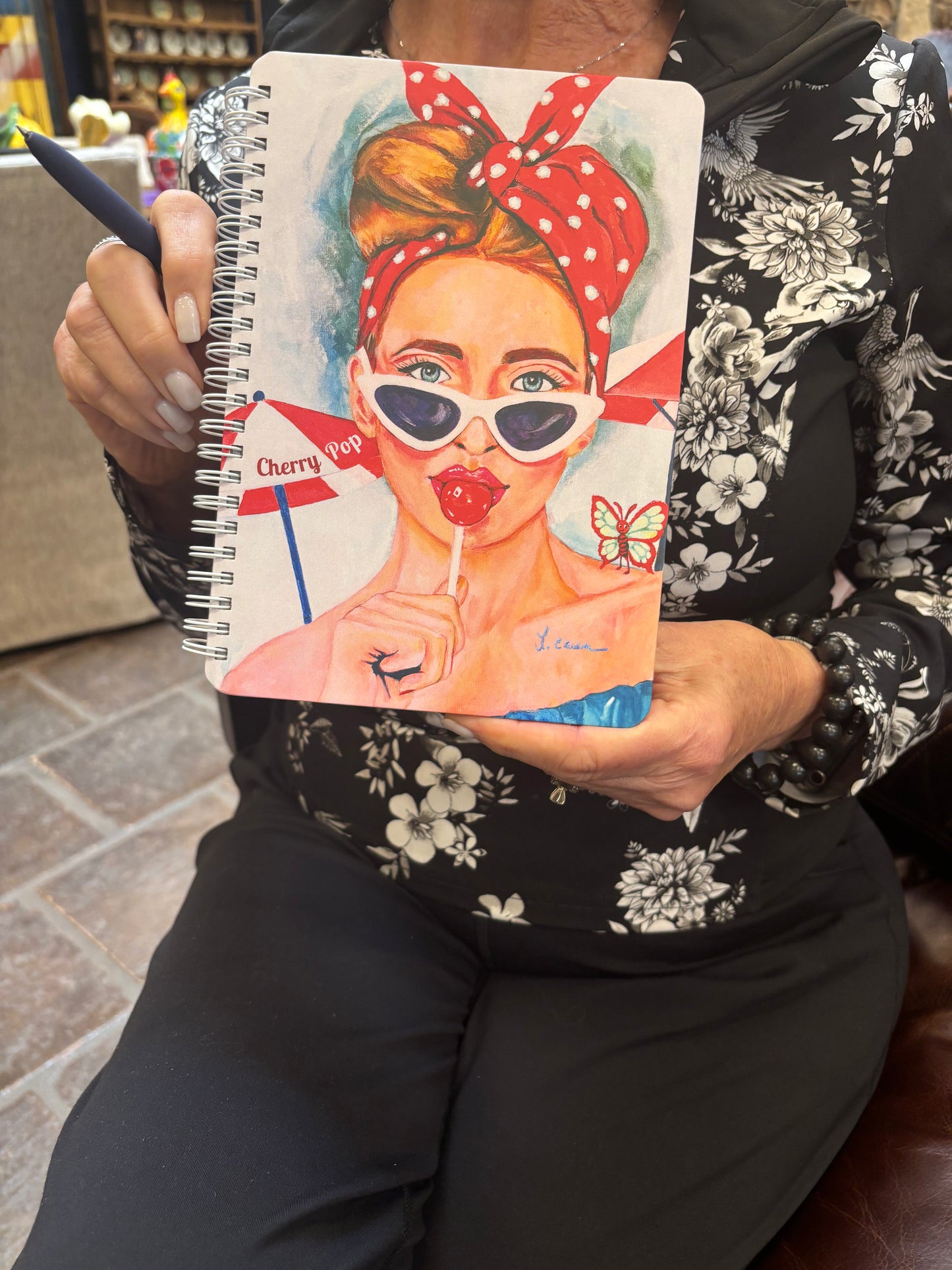 Person holding the Cherry Pop art notebook featuring a retro red-haired girl with a lollipop; lifestyle photo highlighting the notebook’s colorful 60s Charmers design.
