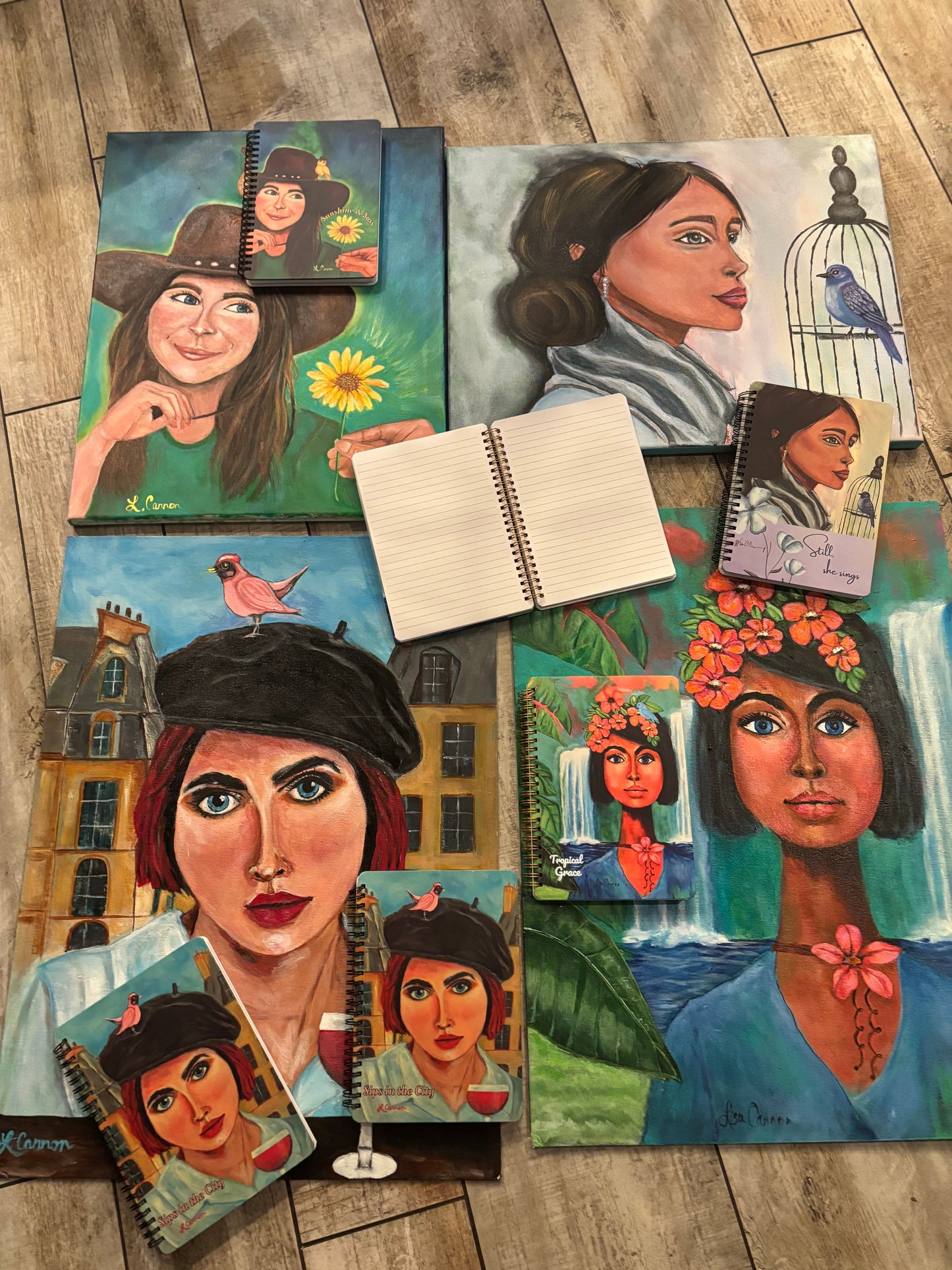 All four original Femmes & Feathers paintings shown with their matching art notebooks resting on each canvas, with an open lined notebook in the center; colorful feminine illustrated journal collection inspired by original artwork.