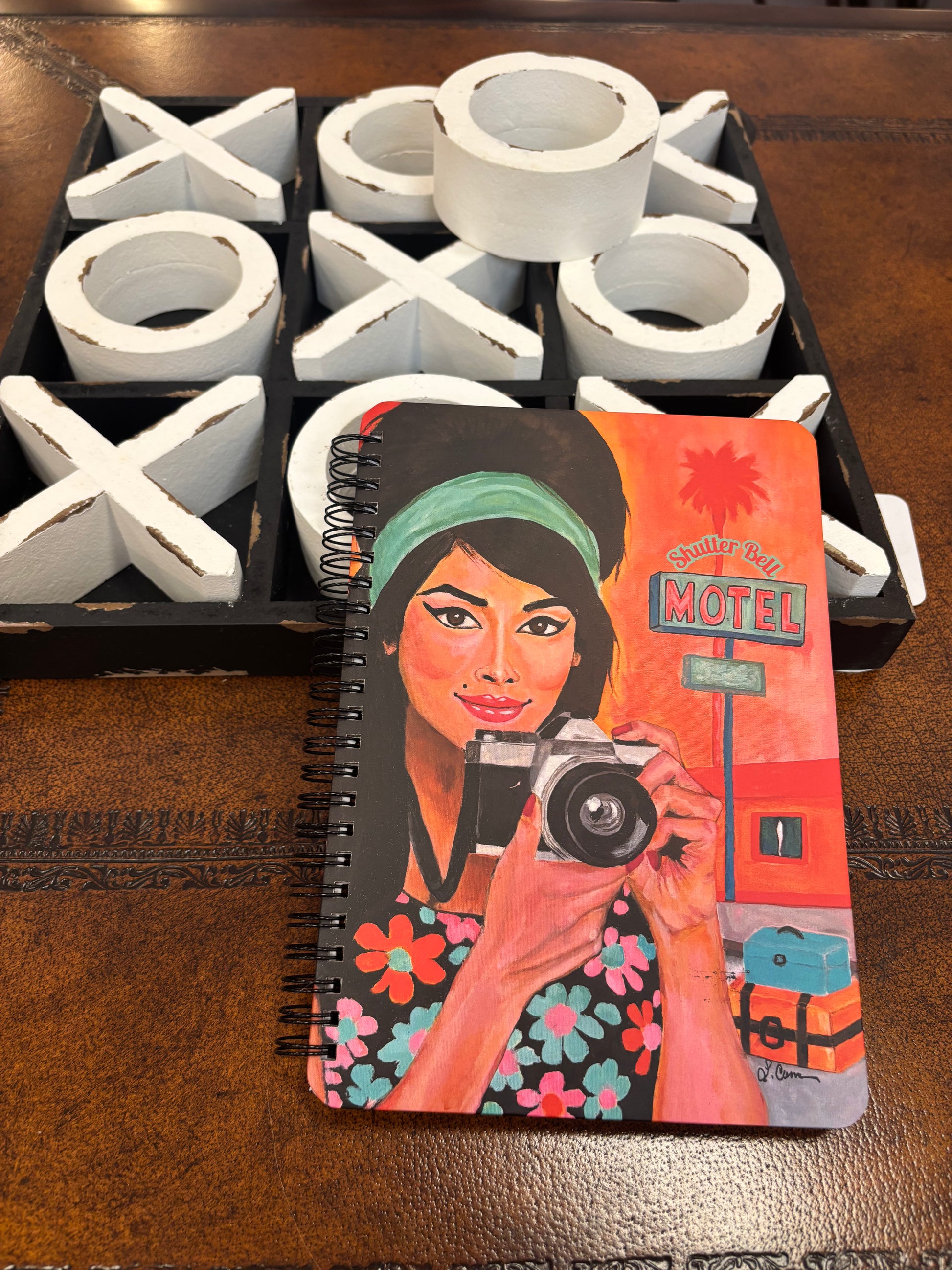 Shutter Bell art notebook featuring a retro camera girl illustration, resting on a tic-tac-toe game board; styled aesthetic notebook photo for product display.