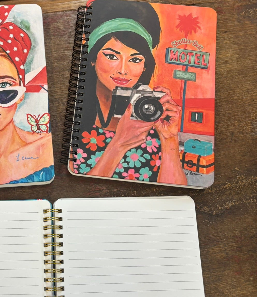 Shutter Bell art notebook front cover with a retro-inspired brunette holding a camera, displayed above an open lined journal; aesthetic notebook photo showing both cover design and interior pages.