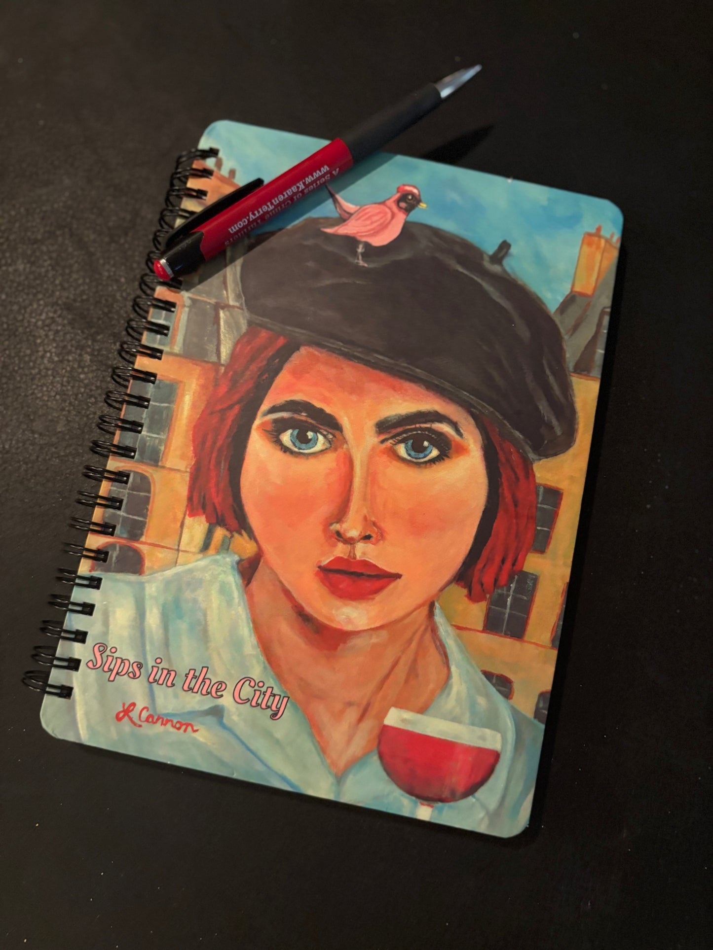Sips in the City art notebook featuring a stylish woman in a beret and a glass of wine, with a pen placed across the top; aesthetic feminine journal from the Femmes & Feathers collection.