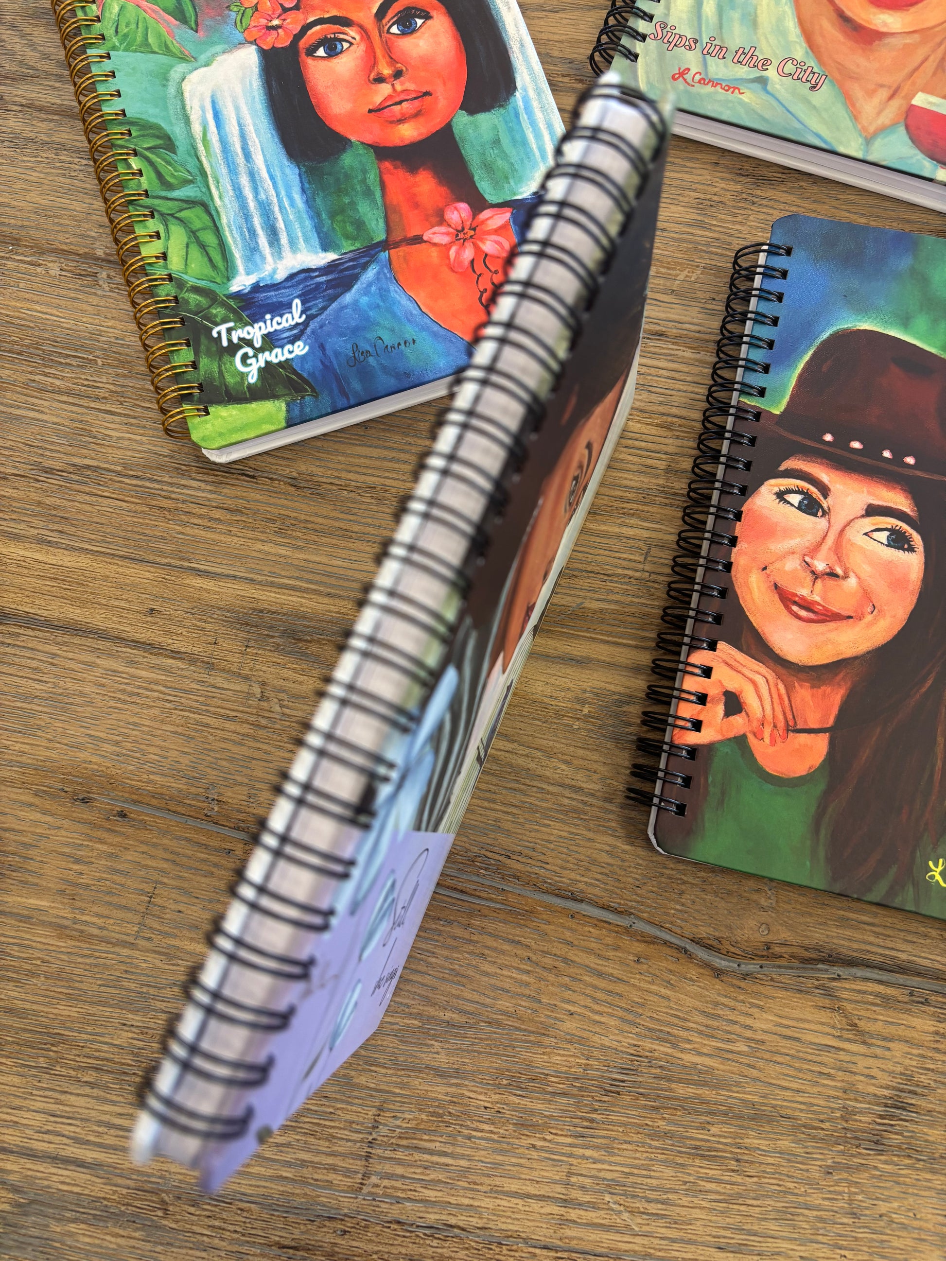 Close-up of a notebook spine with the Tropical Grace and Sunshine & Sass art notebook covers beside it; feminine illustrated journals from the Femmes & Feathers collection displayed together.
