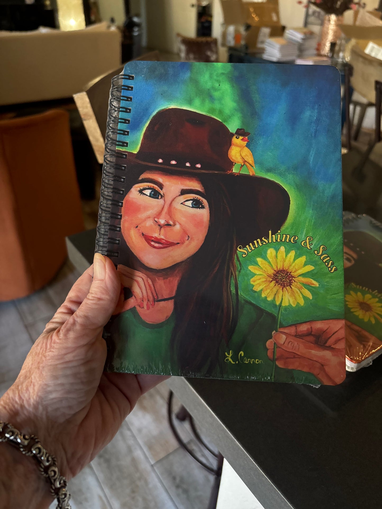 Person holding the Sunshine & Sass art notebook featuring a smiling woman with a yellow bird and sunflower; feminine illustrated journal from the Femmes & Feathers collection shown in a lifestyle photo.