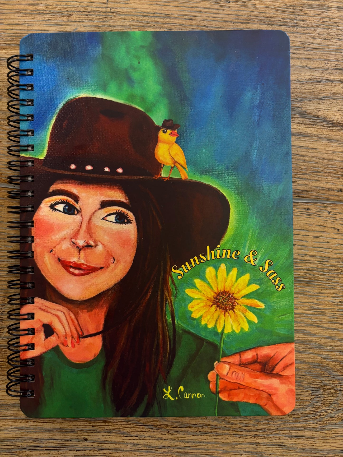 Sunshine & Sass art notebook featuring a smiling woman with a yellow bird and sunflower, displayed on a wood table; colorful feminine illustration from the Femmes & Feathers collection.